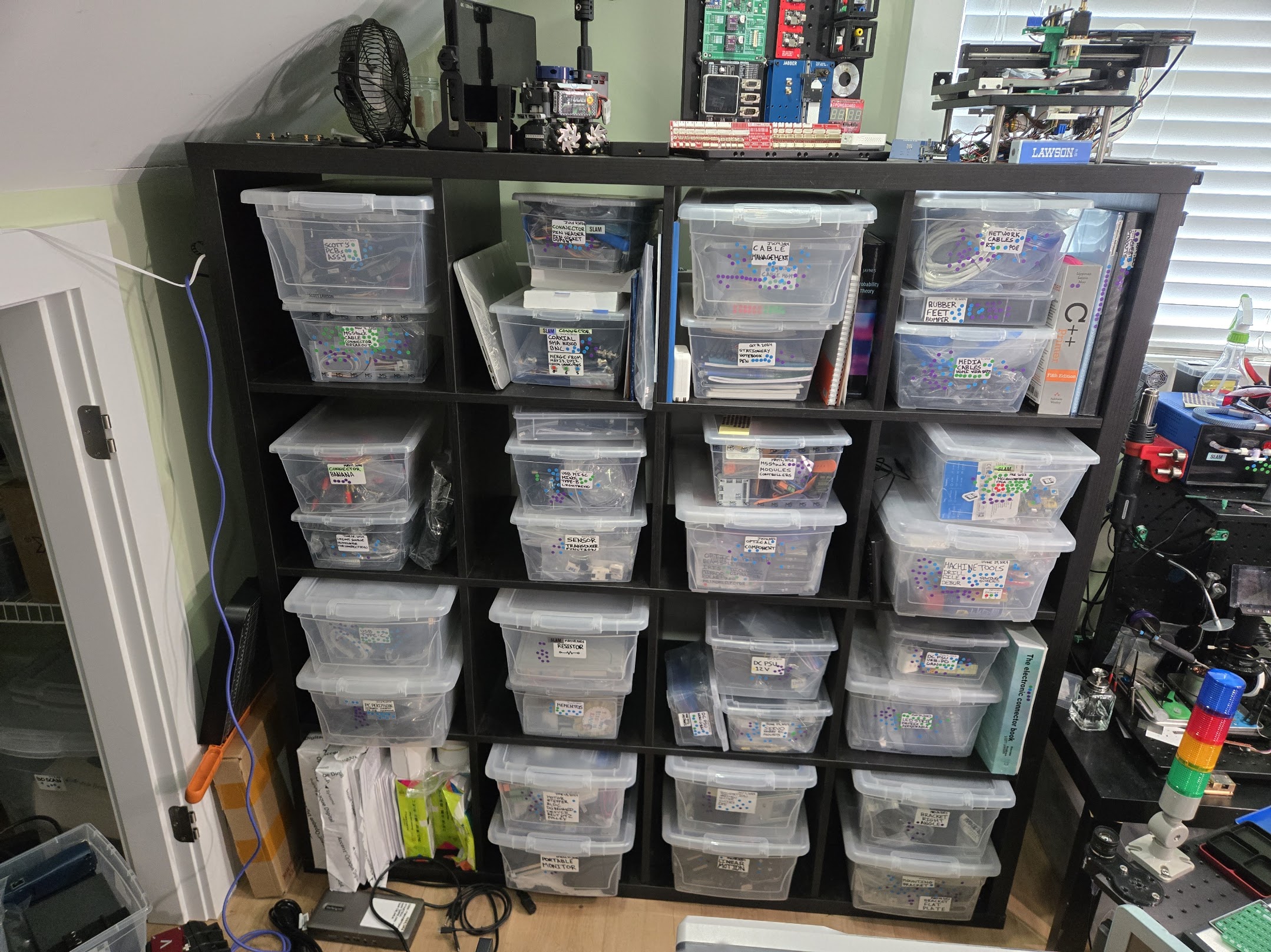 A shelf full of clear boxes in a home lab, each labeled, dated, and covered in colored dot stickers. Some boxes have many dots, others have few. Past projects and PCB designs sit on top.