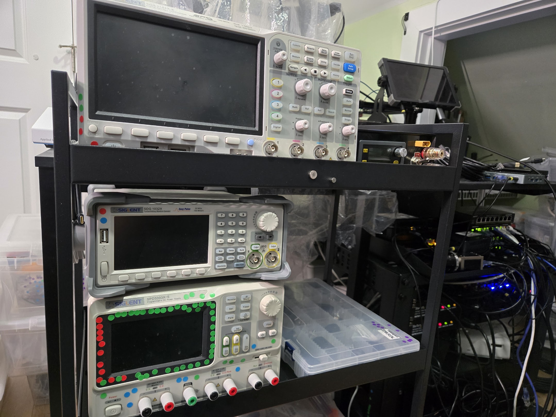 An equipment cart with an oscilloscope, function generator, and Siglent power supply. The power supply is covered in dots while the oscilloscope has five and the function generator has two. A server rack is partially visible on the right.