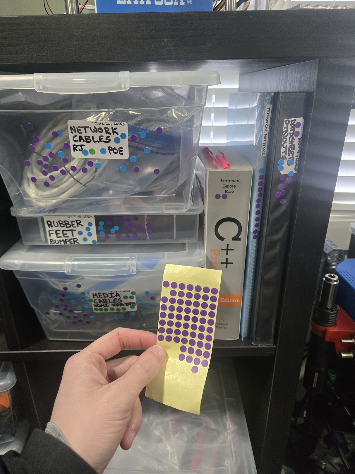A hand holding a sheet of purple dot stickers in front of storage bins for network cables, rubber feet, and media cables. A C++ reference book and technical reference binders are visible with dots on them too.