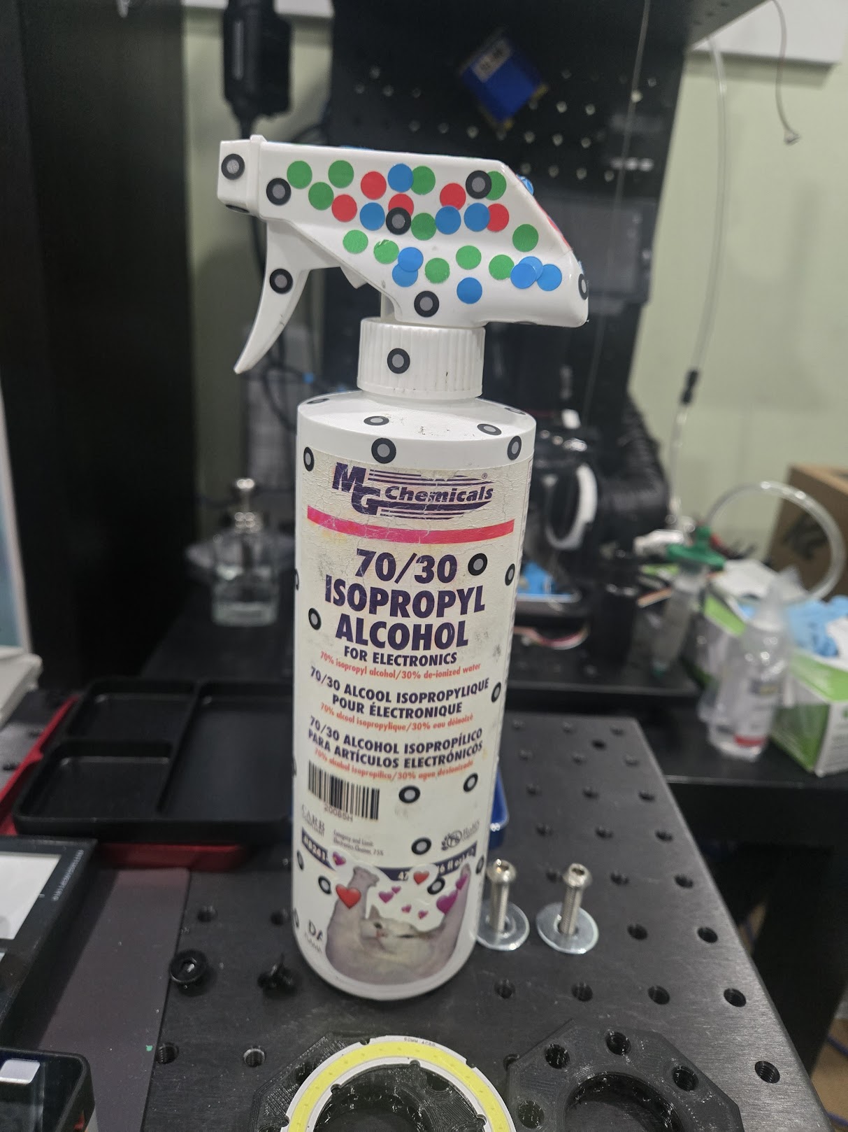 An isopropyl alcohol spray bottle covered in red, green, and blue dot stickers from multiple years of use, with a soldering workstation visible in the background.